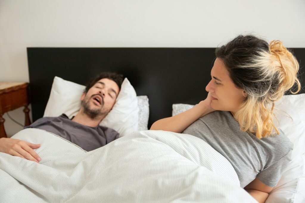 Casal deitado na cama, com o homem dormindo e a mulher acordada olhando ele.